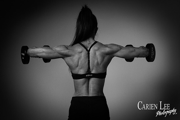 Black-and-white rear fitness portrait of a woman holding dumbbells with arms extended.